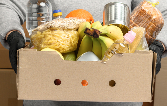 person holding a box of food containing, water, bananas, pasta, bread and other staple items.