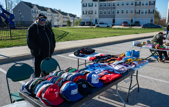 Man standing behind his table at the yard sale. Table is full of baseball caps and other items. 