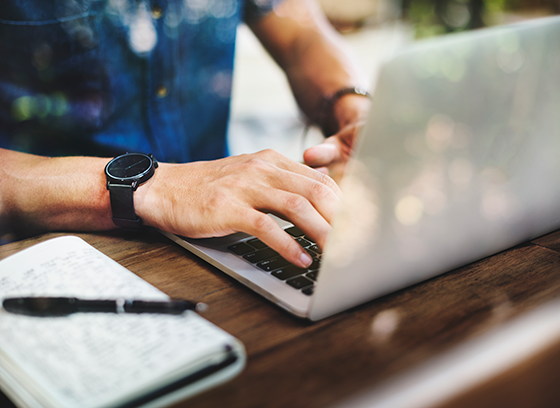 A person typing something on a laptop, with a notepad next to them