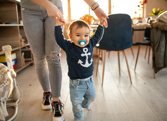 Toddler with pacifier in mouth walking by holding hands with adult, of whom you only see lower half of body