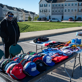 Man standing behind his table at the yard sale. Table is full of baseball caps and other items. 